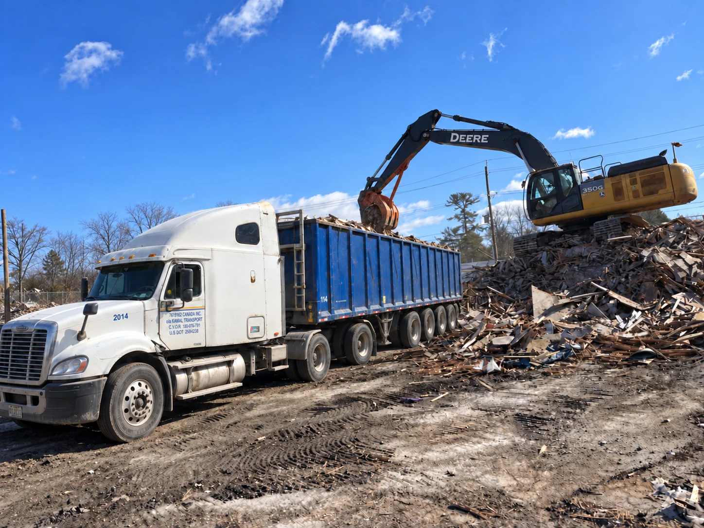 An excavator loading wood debris into a Kamal Transport blue trailer
